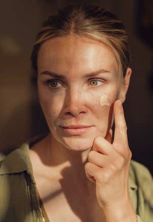 Eine Frau trägt eine leichte Creme auf ihre Wange auf. Sie hat helle Haut mit Sommersprossen, grüne Augen und trägt ein grünes Hemd mit Knöpfen.