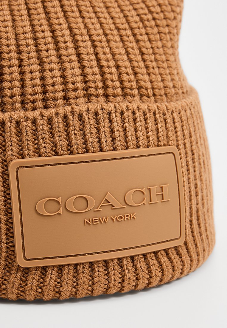 Brun stickad mössa med ribbad textur, som har en rektangulär, beige l�äderpatch prydd med "COACH NEW YORK".