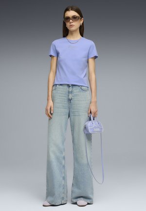 Kobieta ubrana w jasnofioletową koszulkę Puma, jasnoniebieskie jeansy o szerokich nogawkach, okulary przeciwsłoneczne, trzymającą małą dopasowaną fioletową torebkę Puma.