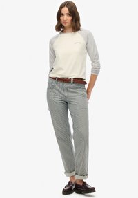 Vrouw staande, draagend een crèmekleurige top met grijze mouwen, gestreepte opgerolde broek, bruine gevlochten riem en donkerbruine loafers tegen een witte achtergrond.