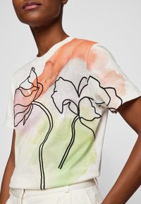 Camiseta de algodón blanca con un diseño floral con contornos negros, acentos en acuarela naranja y verde, y un pequeño logotipo centrado debajo del escote.