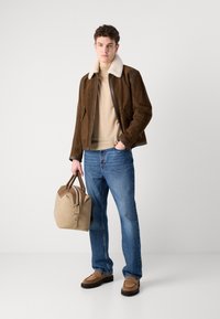 Brun semsket skinnjakke med hvit pelskrage, beige genser, blå jeans og brune mokkasiner. Holder en sandfarget duffelbag med lærdetaljer.