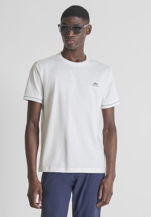 Giovane uomo con occhiali da sole scuri, t-shirt bianca a maniche corte con piccolo logo e pantaloni blu navy su sfondo semplice.