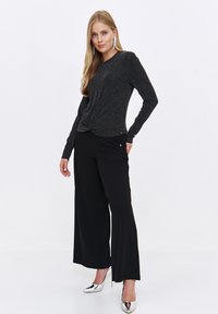 Czarny longsleeve z fakturowanym wykończeniem, z wiązanym przodem. Połączony z szerokimi czarnymi spodniami i metalicznymi szpilkami.
