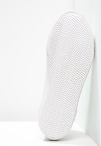 Biała podeszwa trampka z falistym wzorem zapewniającym przyczepność, gładkie krawędzie oraz teksturowana górna część przechodząca w jednolitą białą powierzchnię.