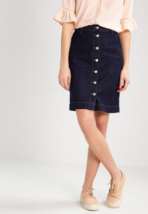 Frau trägt einen dunklen Jeansrock mit Knopfleiste vorne, eine hellrosa Bluse mit Rüschenärmeln und hellrosa Turnschuhe, steht mit überschlagenen Beinen.