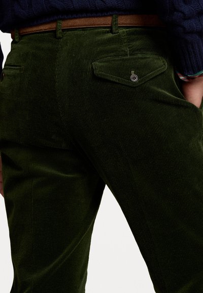 Grønne fløyelsbukser har en slim passform, vertikal ribbet tekstur og to baklommer med trykknapper. En brun belte er synlig.