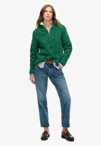 Suéter verde de punto trenzado con media cremallera, combinado con jeans de mezclilla azul relajados y mocasines marrones. Presenta una camisa de rayas azules en capas debajo.