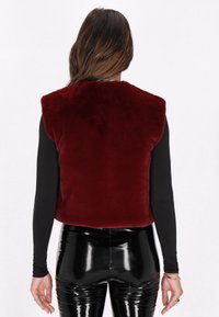 Bordeaux nepbontvest zonder mouwen, met ronde schoudernaden, gecombineerd met zwarte glanzende vinylbroek en een zwart T-shirt met lange mouwen.