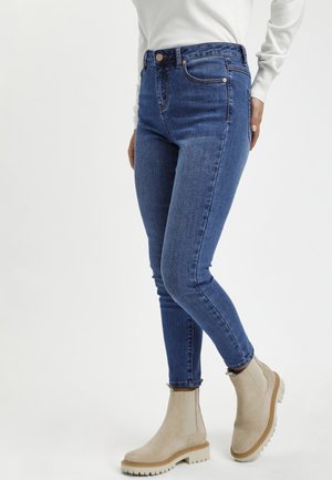 Personne portant un jean skinny bleu et des bottines beiges, debout devant un fond clair uni.