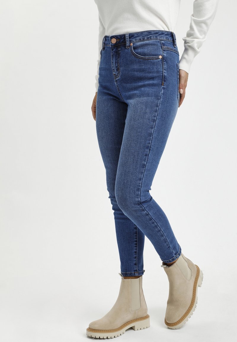 Personne portant un jean skinny bleu et des bottines beiges, debout devant un fond clair uni.
