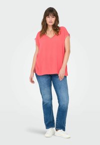 Top rosa corallo ampio con scollatura a V e bordo in pizzo ai lati delle maniche, abbinato a jeans dritti azzurri chiari e sneakers bianche.