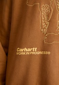 Sudadera de algodón marrón con un diseño bordado y el texto "Carhartt WORK IN PROGRESS" en dorado. Textura suave y ajuste relajado.