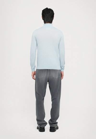 Lyseblå strikkegenser med høy krage, sammen med mørkegrå løstsittende jeans og svarte sko, sett fra baksiden.