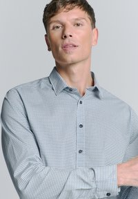 Chemise à boutons à motif avec un design géométrique bleu clair et blanc, avec un col structuré et des boutons bleus, fabriquée en tissu lisse.