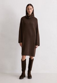 Braunes Kleid mit Schalkragen und langen Ärmeln, aus weichem Strickstoff gefertigt, kombiniert mit knielangen braunen Wildlederstiefeln mit einer robusten Sohle.