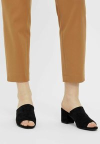 Black suede open-toe mules with a wide strap and a chunky heel, paired with tan ankle-length trousers.