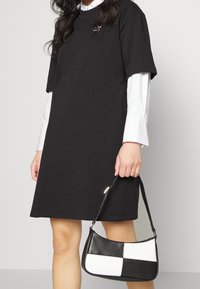 Robe t-shirt noire à manches courtes, portée par-dessus une chemise blanche. Tient un petit sac à main noir et blanc de forme courbée avec une sangle réglable.
