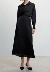 Longue robe noire en tissu lisse, avec un col boutonné, des manches longues et un détail de taille torsadée. Portée avec des talons noirs pointus.