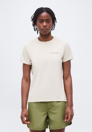 Žena s krátkymi dredmi, oblečená v béžovom tričku Norrona a olivovo zelených šortkách, stojaca na svetlom jednoliatom pozadí.