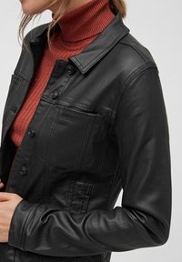 Chaqueta de cuero negra con un diseño de cuello, que presenta cierres de botón y un bolsillo en el pecho. Lleva puesta una camiseta de cuello alto de color óxido con textura acanalada.