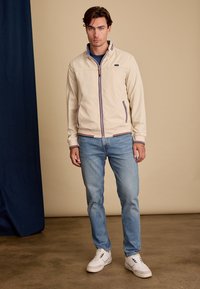 Veste légère beige avec fermeture éclair sur le devant, col contrasté et accents rayés. Associée à un jean bleu et des baskets blanches.