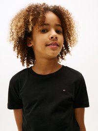 T-shirt en coton noir à manches courtes, avec un petit logo rouge, blanc et bleu sur la poitrine gauche. Coiffure bouclée.