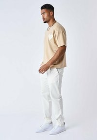 Camiseta de manga corta beige con un diseño de corazón en el lado izquierdo del pecho, combinada con pantalones blancos y zapatillas blancas. Afit simple y relajado.
