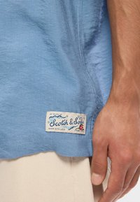Ανοιχτό μπλε πουκάμισο με κοντά μανίκια και απαλή υφή. Διαθέτει ραμμένη ετικέτα που αναγράφει "Scotch & Soda" με διακοσμητικές λεπτομέρειες.