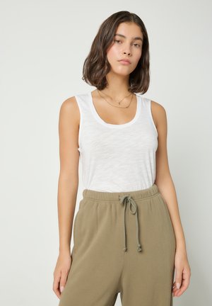 Noor naine õlakõrguse pruunide juustega, kes kannab valget varrukateta t-särki ja oliivirohelisi tõmblukuga pükse, seisab ühevärvilise tausta ees.