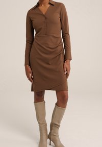 Brun långärmad klänning med krage, med tre knappar och rynkad längd vid midjan, i kombination med beige knähöga stövlar.