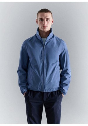 Joven con el pelo corto que lleva una chaqueta azul con cremallera y pantalones oscuros, de pie con las manos en los bolsillos sobre un fondo liso.