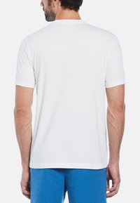 Witte katoenen t-shirt met korte mouwen, ronde hals en een gladde textuur; gedragen met teal gekleurde shorts met textuur, waarbij de achterkant zichtbaar is.