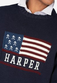 Granatowy sweter z dzianiny z niebieską i czerwoną łatą przedstawiającą wzór flagi oraz napisem "HARPER" białymi literami poniżej emblematu. Posiada ściągacz przy kołnierzyku.