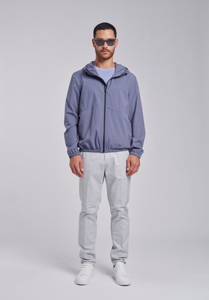 Mand iført solbriller, en lys lilla lynlåstrøje med hætte, lysegrå bukser og hvide sneakers, stående mod en ensfarvet baggrund.