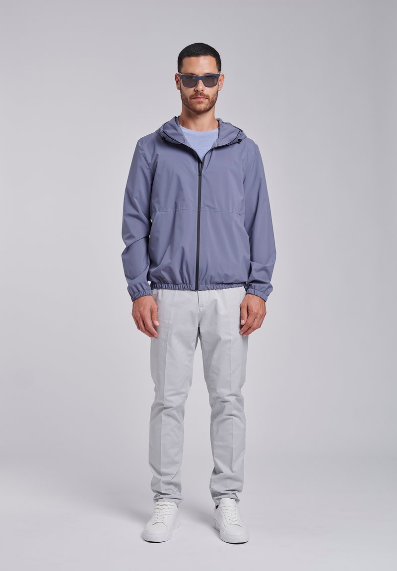 Uomo con occhiali da sole, giacca con cappuccio viola chiaro con cerniera, pantaloni grigio chiaro e scarpe da ginnastica bianche, in piedi davanti a uno sfondo semplice.