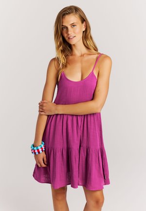 Vrouw met lang blond haar, gekleed in een mouwloze paarse jurk en kleurrijke armbanden, staand tegen een effen lichte achtergrond.