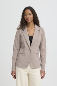Mujer con cabello largo y rizado, vestida con un blazer beige sobre una camiseta negra y pantalones crema, posando frente a un fondo claro y liso.