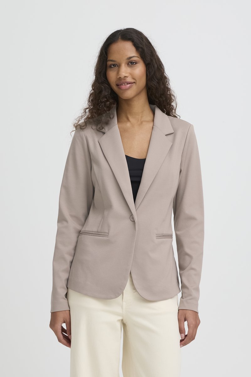Mujer con cabello largo y rizado, vestida con un blazer beige sobre una camiseta negra y pantalones crema, posando frente a un fondo claro y liso.