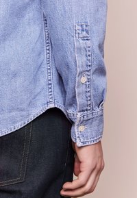 Ljust blå denimskjorta med knappad manschett, med sömnadsdetaljer och en något fransad nederkant. Bärs över mörka jeans.
