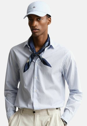 Uomo con cappellino bianco, camicia a righe azzurro chiaro con bottoni, foulard da collo blu navy e pantaloni beige su sfondo semplice.