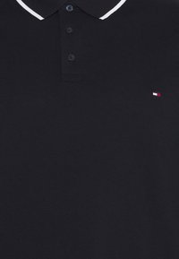 Mustapaita, joka on valmistettu teksturoidusta kankaasta. Siinä on valkoinen raidallinen kaulus, kolmen napin nappilista ja pieni logo punaisessa, valkoisessa ja sinisessä värissä.