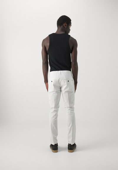 Top senza maniche nero, pantaloni bianchi slim-fit e scarpe nere con suola testurizzata. La vista mostra la parte posteriore dell'outfit su uno sfondo semplice.