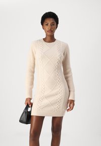 Strikket kjole i lys beige med teksturerte kabelmønstre, lange ermer og ribbestrikkede mansjetter. Modellen holder en liten svart håndveske.
