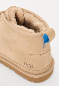 Botín de ante color marrón claro con un suave forro de piel de peluche, acento azul en el talón y suela de goma. Presenta el logotipo "UGG" grabado en el costado.