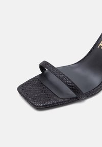 Μαύρο σανδάλι με ψηλό τακούνι, τετράγωνη μύτη και λεπτό υφασμάτινο λουράκι μπροστά.