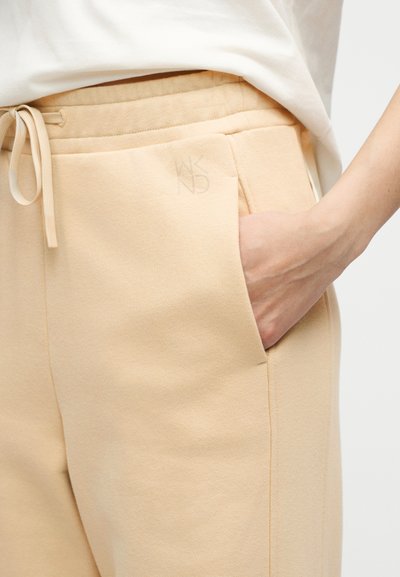 Δεξί χέρι στην τσέπη μπεζ παντελονιού με κορδόνι, φορεμένο με λευκό τοπ, που δείχνει την απαλή υφή του υφάσματος και το διακριτικό λογότυπο στην τσέπη.