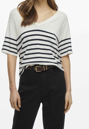 Vrouw draagt een wit T-shirt met korte mouwen en V-hals met marineblauwe horizontale strepen, zwarte broek en een bruine riem met een gouden gesp.