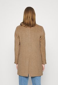 Henkilö, jolla on olkapituinen ruskea hius ja päällään ruskea villakangas takki sekä siniset farkut, seisoo selkä kohti yksinkertaista valkoista taustaa.