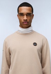 Felpa beige con un patch rotondo sul petto. Design a girocollo, texture morbida, abbinata a un dolcevita bianco sotto.